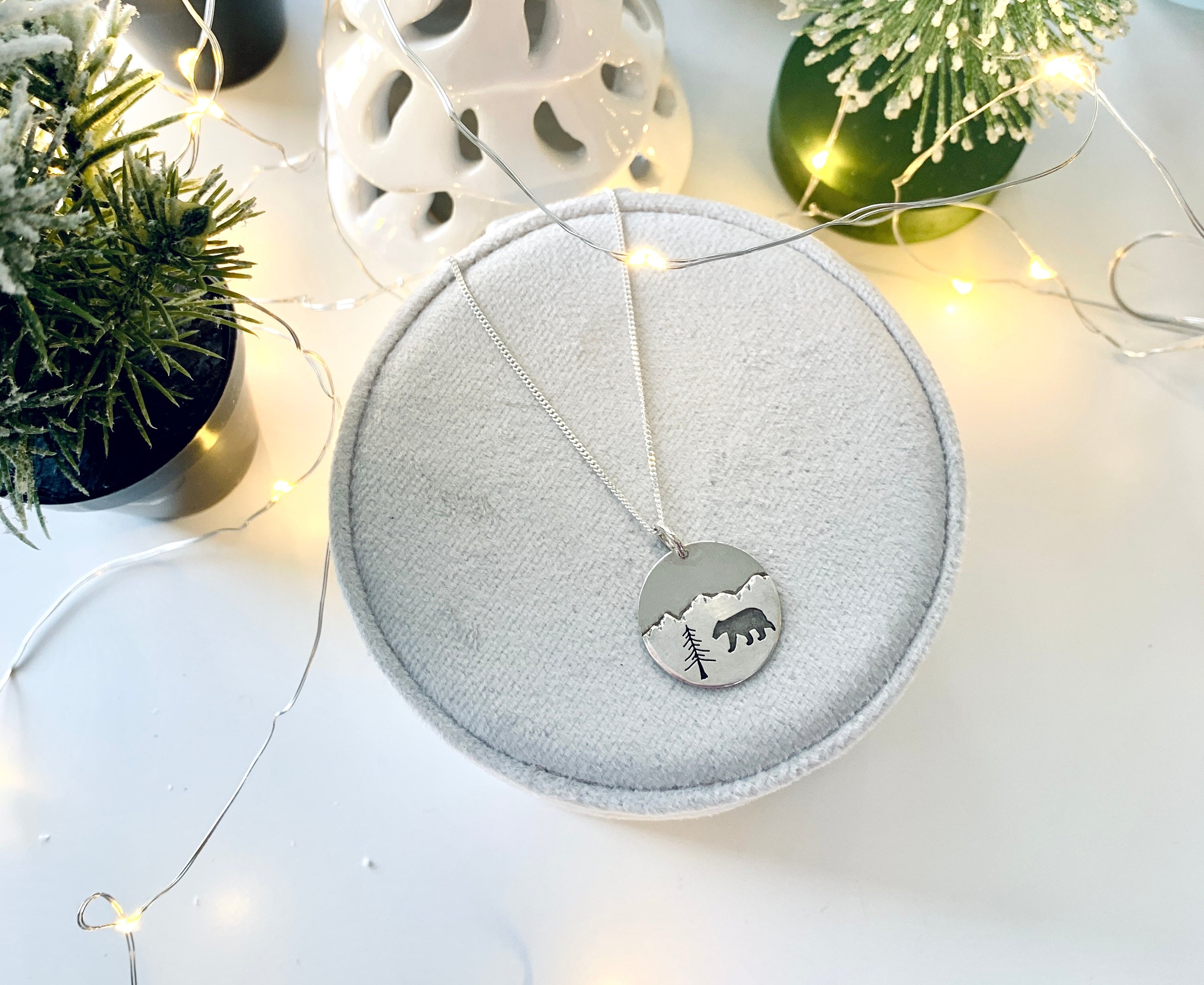 Decorative christmas items including a white ceramic tree, a round gray jewellery case with a bear pendant laying on it, and small green trees on a light surface.