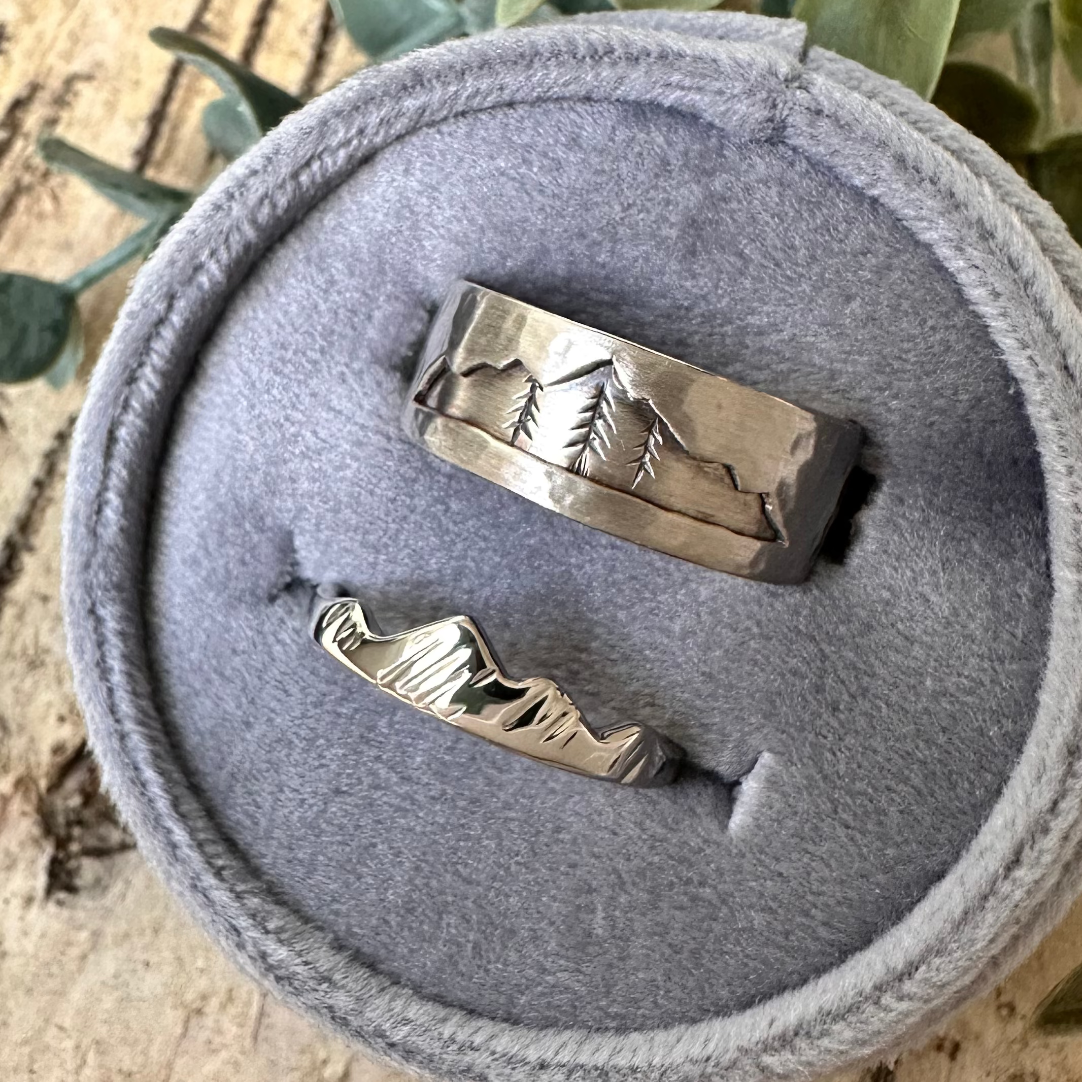 Two silver rings in a gray velvet ring box with green leaves on a wooden surface