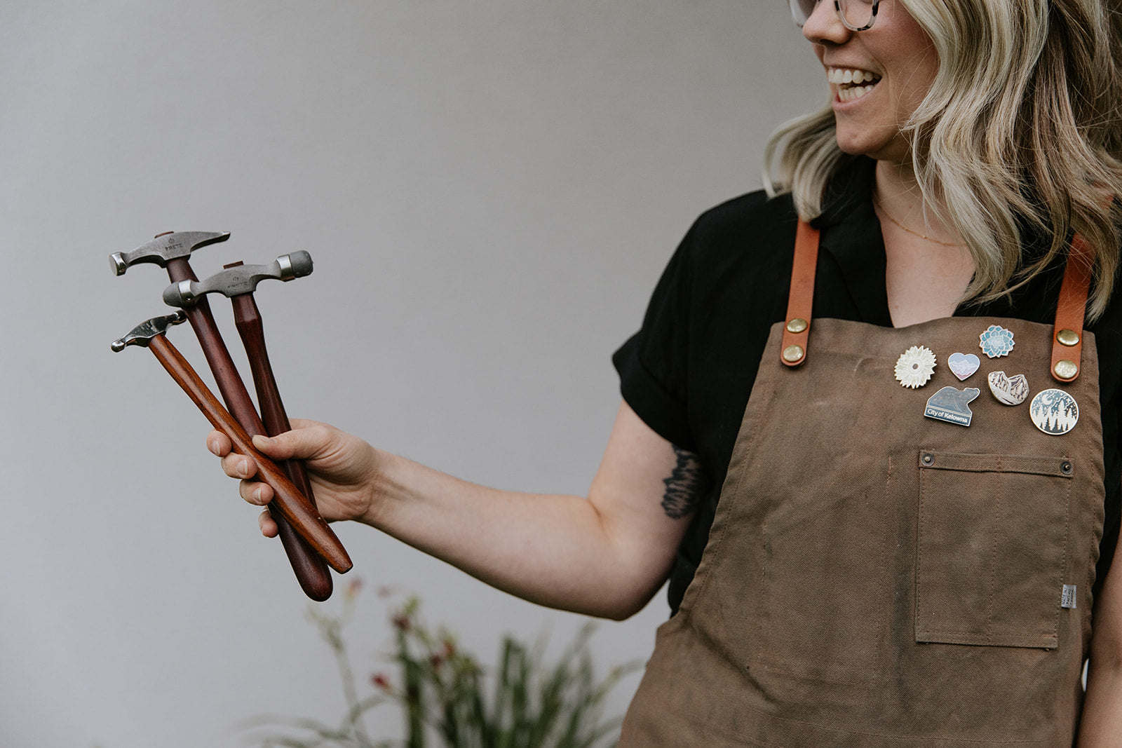 A blond haired mom of twins stands outside showing off her hammers from her goldsmith studio